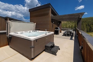 Un bain à remous sur une terrasse extérieure, surmontée d’une pergola en bois, avec un barbecue et une vue sur une colline boisée.