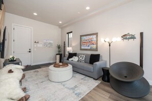 Ein Wohnzimmer mit einem grauen Sofa, einem runden Hocker, einer schwarzen Stehlampe und einem großen schwarzen Lautsprecher.