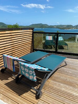 Eine Terrasse mit zwei Liegestühlen, einer mit einem gestreiften Handtuch und der andere mit einem Blumenhandtuch, mit Blick auf eine malerische Bucht und Hügel.