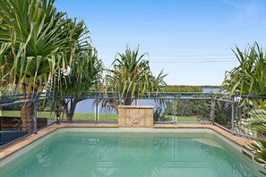 Ein Swimmingpool mit einer Steinfountain, umgeben von Palmen und Blick auf ein Gewässer.