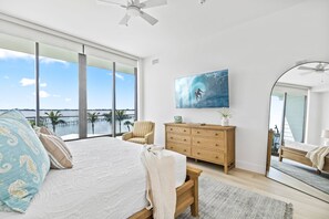 A bedroom with a large bed, a wooden dresser, a chair, and a painting. - Sunset Shores Retreat | 3BR River-View Condo w/ Double Balcony & Private Beach Access (Melbourne Beach)