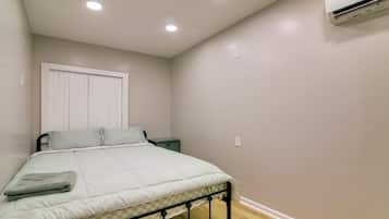 A bedroom with a bed, a nightstand, a window with blinds, and an air conditioning unit.