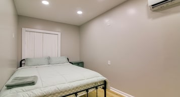 Un dormitorio con cama, mesita de noche, ventana con persianas y equipo de aire acondicionado.