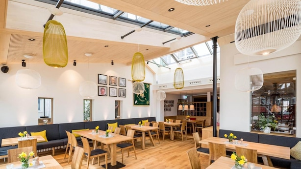 A modern dining area with wooden tables and chairs, pendant lighting, and a high ceiling with skylights. - Waldhorn (Kempten)
