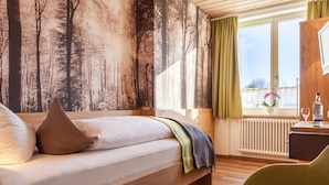 A bedroom with a bed, a window with curtains, a radiator, and a small table with a bottle and glass. - Waldhorn (Kempten)
