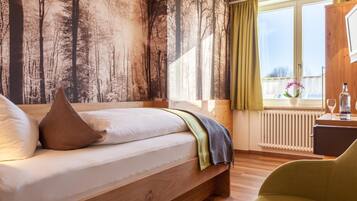 Ein Schlafzimmer mit einem Bett, einem Fenster mit Vorhängen, einem Heizkörper und einem kleinen Tisch mit einer Flasche und einem Glas.