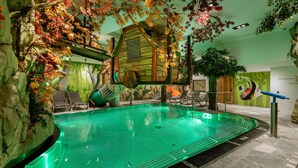 Una piscina interior con una estructura de cabaña de madera, sillones de descanso y follaje artificial.