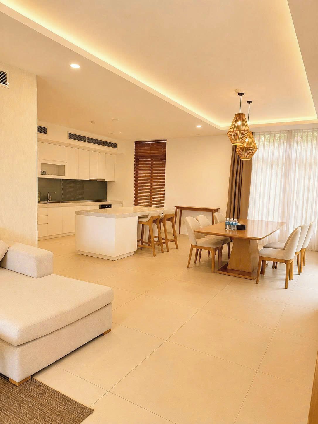 Una sala de estar moderna con un sofá, un comedor con mesa y sillas, una cocina con armarios blancos y un ventanal con persianas.