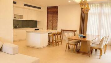 Una sala de estar moderna con un sofá, un comedor con mesa y sillas, una cocina con armarios blancos y un ventanal con persianas.