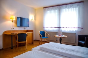 Quarto de hotel com cama, escrivaninha, cadeira, televisão e janela com cortinas.