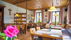 Een restaurant met houten tafels en banken, een kruis aan de muur en een raam met gordijnen.