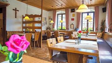 A restaurant with wooden tables and benches, a cross on the wall, and a window with curtains.
