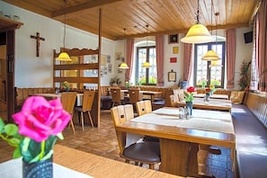 Restaurante com mesas e bancos de madeira, uma cruz na parede e uma janela com cortinas.