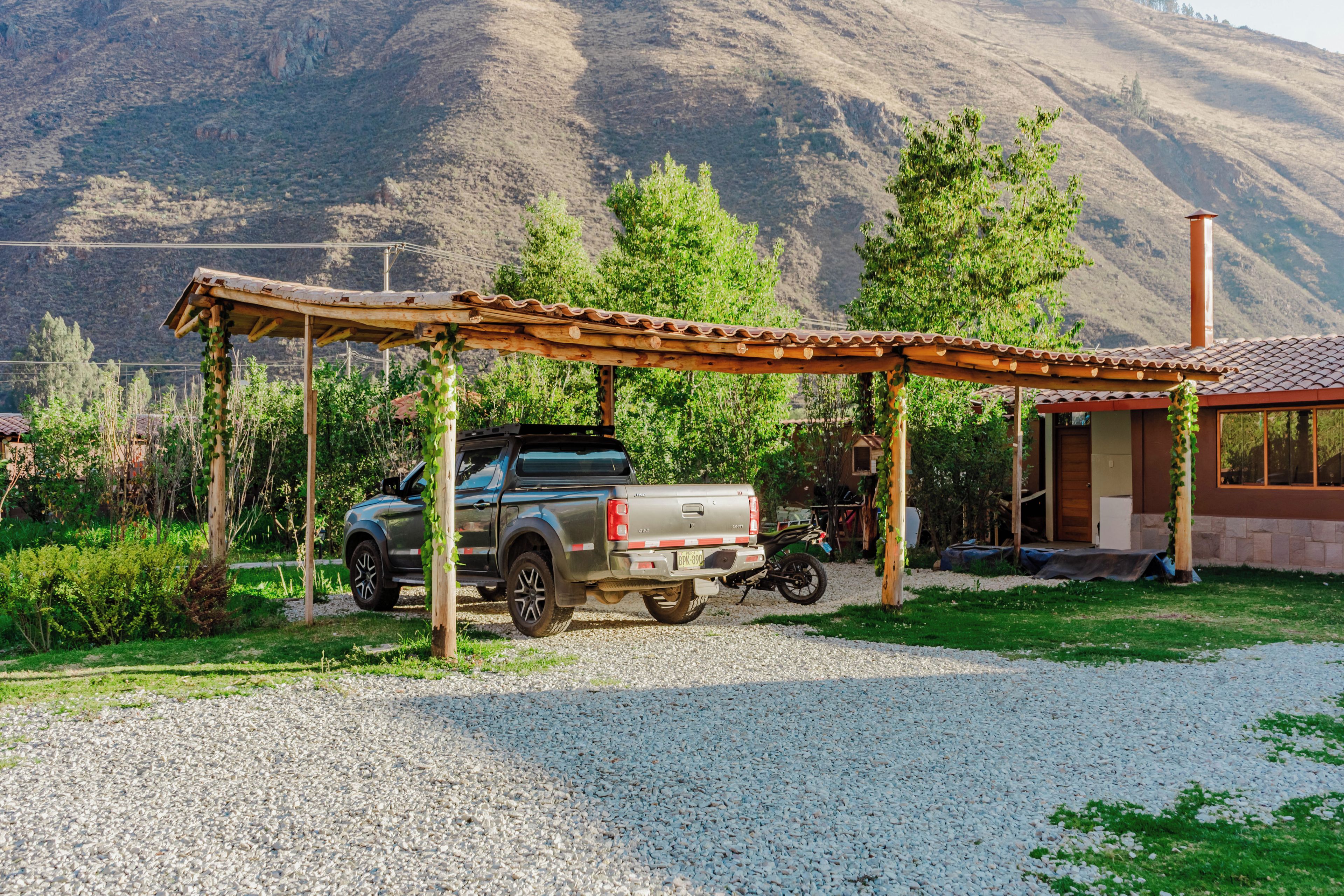 A gravel driveway leading to a wooden structure with a metal roof, a pickup truck parked under it, and a mountainous backdrop.