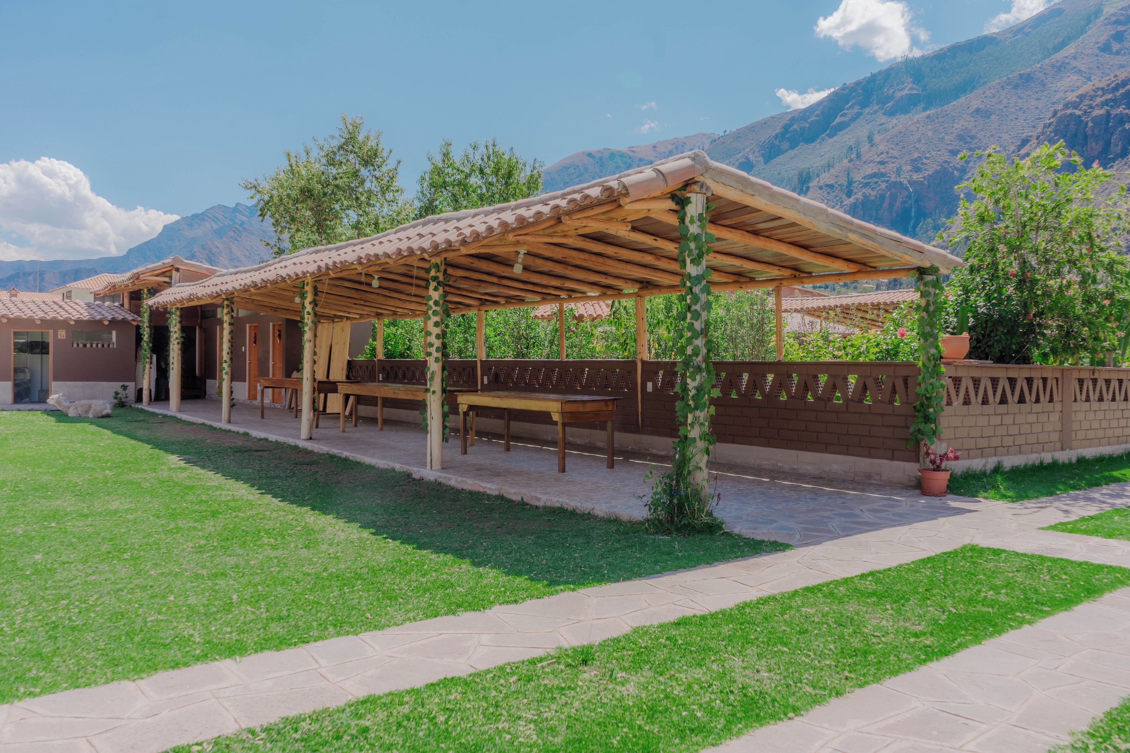 Un espacio al aire libre techado con mesas y sillas, un muro de ladrillo y montañas al fondo.