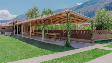 Un espacio al aire libre techado con mesas y sillas, un muro de ladrillo y montañas al fondo.