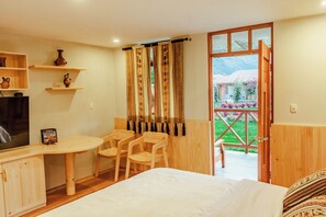 A room with a bed, a desk with chairs, a television, and a window with curtains.
