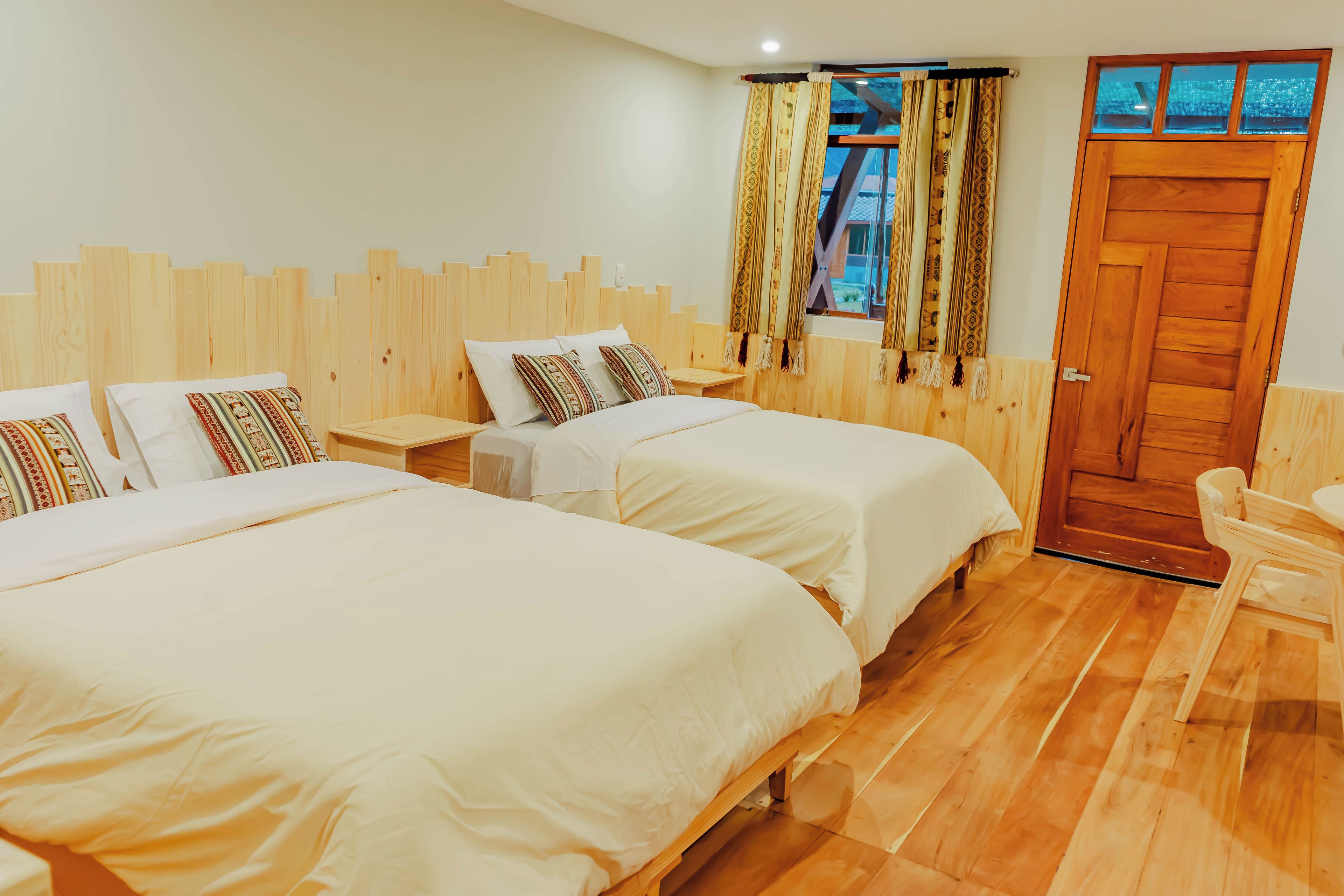 Ett hotellrum med två sängar, en sänggavel i trä, ett fönster med mönstrade gardiner och en dörr i trä.