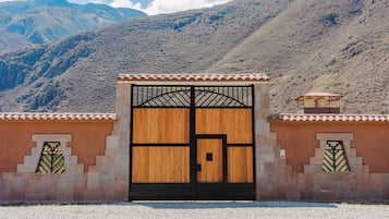 Um grande portão de madeira com arco de metal, ladeado por muros de pedra e um cenário montanhoso.