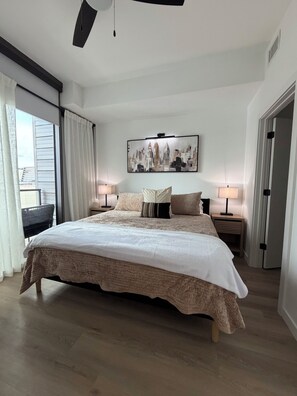A bedroom with a large bed, two bedside tables with lamps, a cityscape painting on the wall, and a window with curtains. - Cozy 1-bedroom apartment in charming Atlanta with AC and gym access (Atlanta)