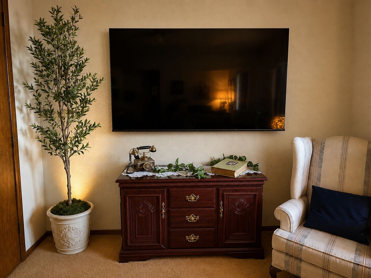 A living room with a television mounted on the wall, a wooden cabinet with drawers, a potted plant, and a striped armchair.