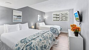 A hotel room with two beds, a TV, a desk with a flower vase, and a window with a view of buildings. - Oceanfront Luxury | 2BR Condo| No Resort Fees (Hollywood)