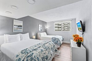 Ein Hotelzimmer mit zwei Betten, einem Fernseher, einem Schreibtisch mit Blumenvase und einem Fenster mit Blick auf Gebäude.