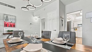 A dining area with a long table set for four, featuring a modern chandelier, white walls, and a view into the kitchen. - Oceanfront Luxury | 2BR Condo| No Resort Fees (Hollywood)