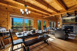A wooden cabin interior with a dining area, a fireplace, and large windows. - Mountain Views, Four King Bedrooms, Hot Tub, Pool, Game Room &( EV Charger) (Pigeon Forge)