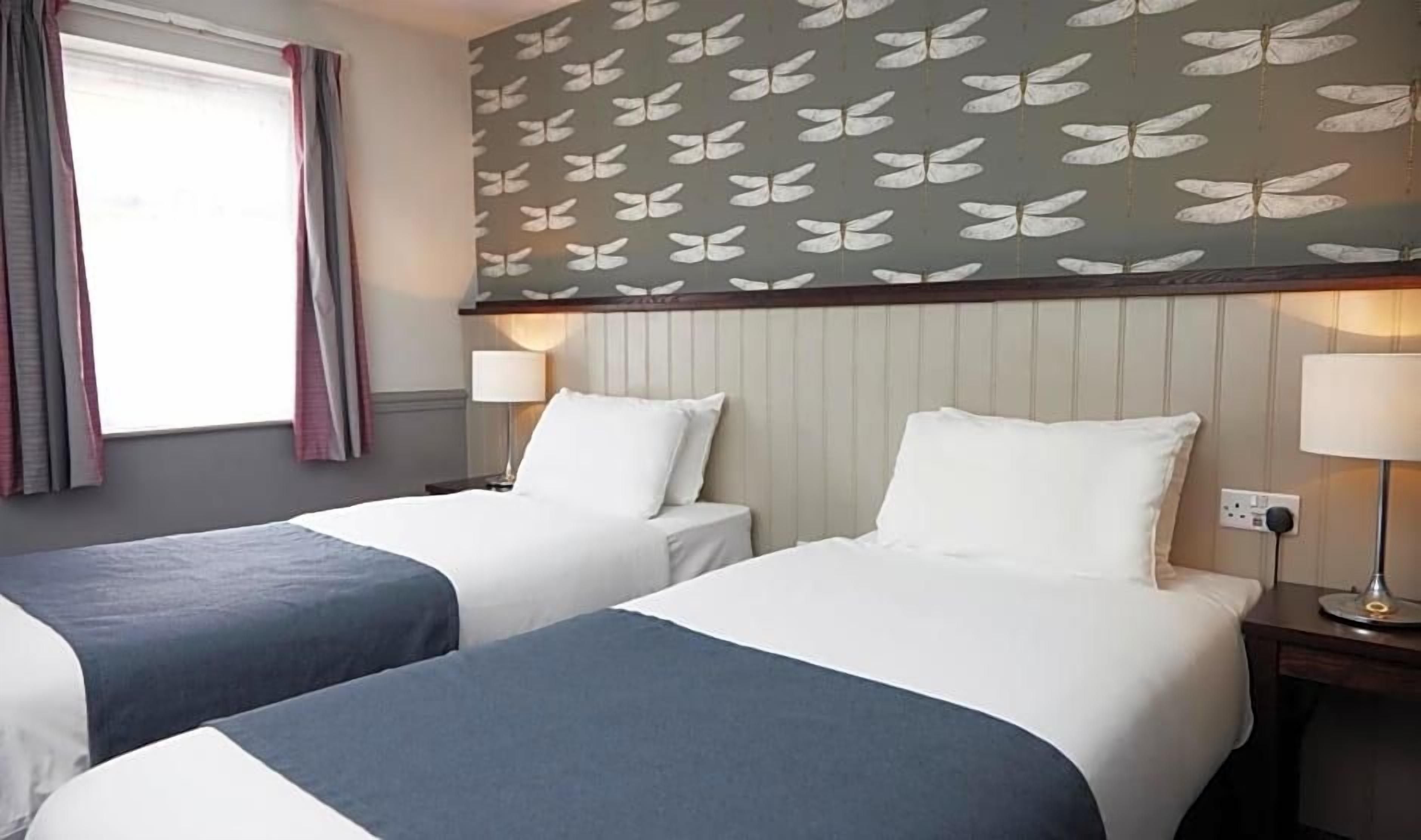 A hotel room with two beds, a window with curtains, a wall with a butterfly pattern, and bedside lamps.