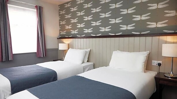 A hotel room with two beds, a window with curtains, a wall with a butterfly pattern, and bedside lamps. - Charnwood Arms by Good Night Inns (Coalville)