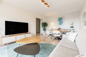 Una sala de estar moderna con un televisor de pantalla plana, una mesa de centro de madera, una silla negra, un sofá blanco y una zona de comedor con mesa y sillas.