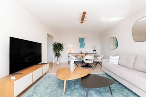 Ein modernes Wohnzimmer mit einem Flachbildfernseher, einer Holzkommode, einem runden Couchtisch, einem grauen Sofa und einem Essbereich mit Tisch und Stühlen.