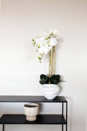 Une orchidée blanche dans un pot blanc, posée sur une étagère noire, avec une tasse en céramique à côté.