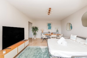 Ein modernes Hotelzimmer mit Flachbildfernseher, einem Holznachttisch, einem Schreibtisch mit Stuhl, einem Esstisch mit Stühlen, einem Bett mit weißer Bettwäsche, einem grünen Teppich, einer Topfpflanze und einer Deckenleuchte.