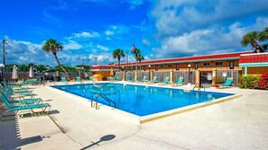Ein Schwimmbecken mit Liegestühlen und Sonnenschirmen, ein Gebäude mit rotem Dach und Palmen im Hintergrund.