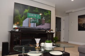 A modern living room with a flat-screen TV mounted on a dark wooden cabinet, a glass coffee table with decorative items, and a cityscape painting on the wall. - Comfort Home (St. John's)