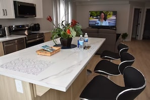A modern kitchen with a large island, black chairs, a flat-screen TV, and a potted plant. - Comfort Home (St. John's)