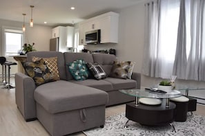 A modern living room with a grey sectional sofa, a glass coffee table, and a kitchen in the background. - Comfort Home (St. John's)