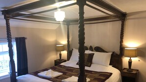 A four-poster bed with white linens and a brown throw, flanked by two bedside tables with lamps, in a room with a window and a chandelier. - The Walnuts Country House (Bury St Edmunds)