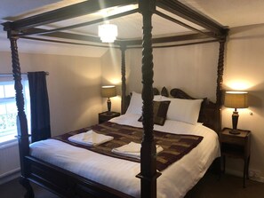 Una cama con dosel, con ropa de cama blanca y un plaid marrón, flanqueada por dos mesitas de noche con lámparas, en una habitación con ventana y candelabro.