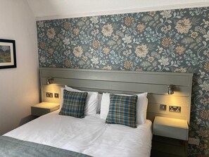 A neatly made bed with white linens and plaid pillows, flanked by bedside tables with lamps, against a floral wallpaper backdrop. - Tullie Inn (Alexandria)