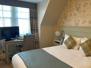 A hotel room with a bed, a desk with a television, a chair, a bedside table with a lamp, and a patterned wall. - Tullie Inn (Alexandria)