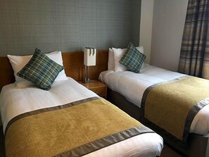 A hotel room with two beds, each with a pillow and a blanket. There is a nightstand between the beds with a lamp on it. - Tullie Inn (Alexandria)