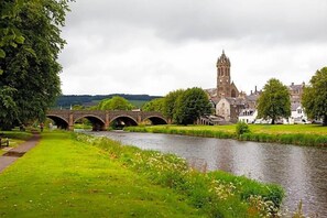 A river with a stone bridge, a church with a tall tower, and a grassy area with trees. - Green Tree Hotel (Peebles)