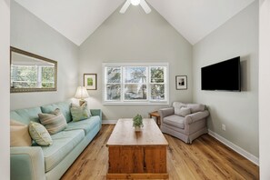Ein Wohnzimmer mit einem hellblauen Sofa, einem karierten Sessel, einem Holztisch und einem an der Wand befestigten Fernseher.
