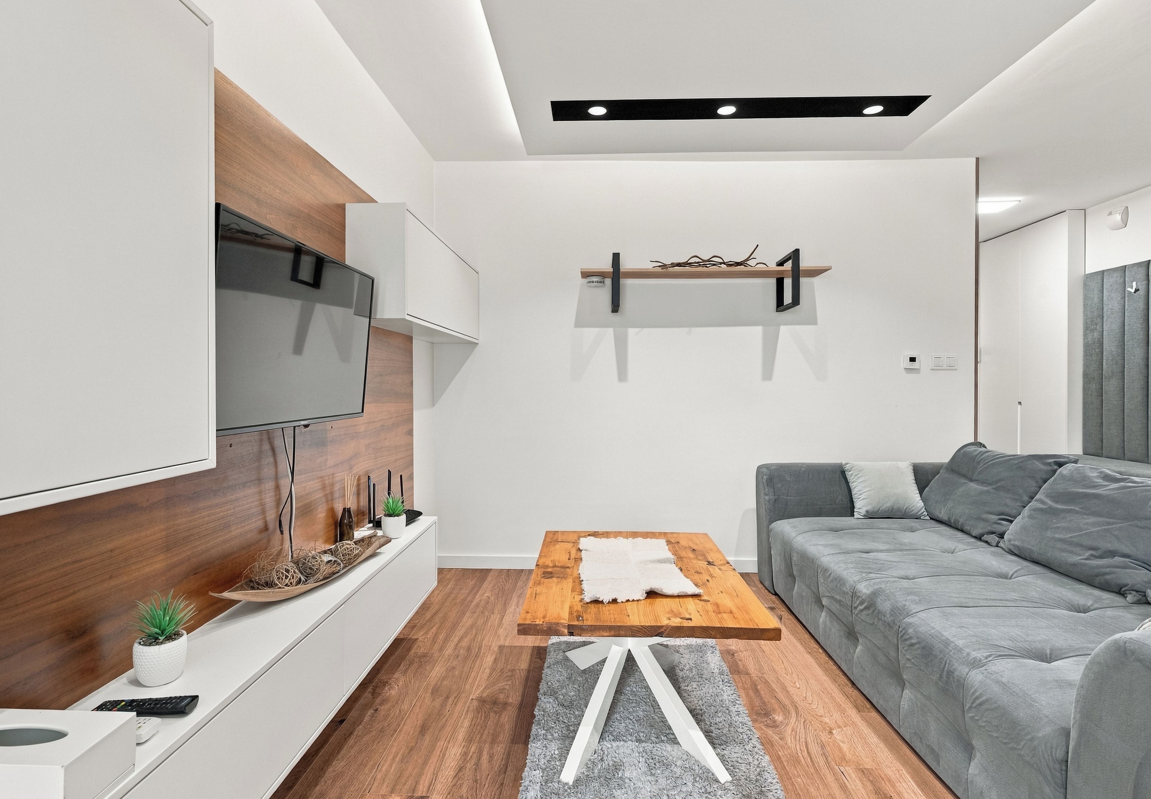 Ein modernes Wohnzimmer mit Flachbildfernseher, Holzboden, grauem Sofa und Couchtisch aus Holz.
