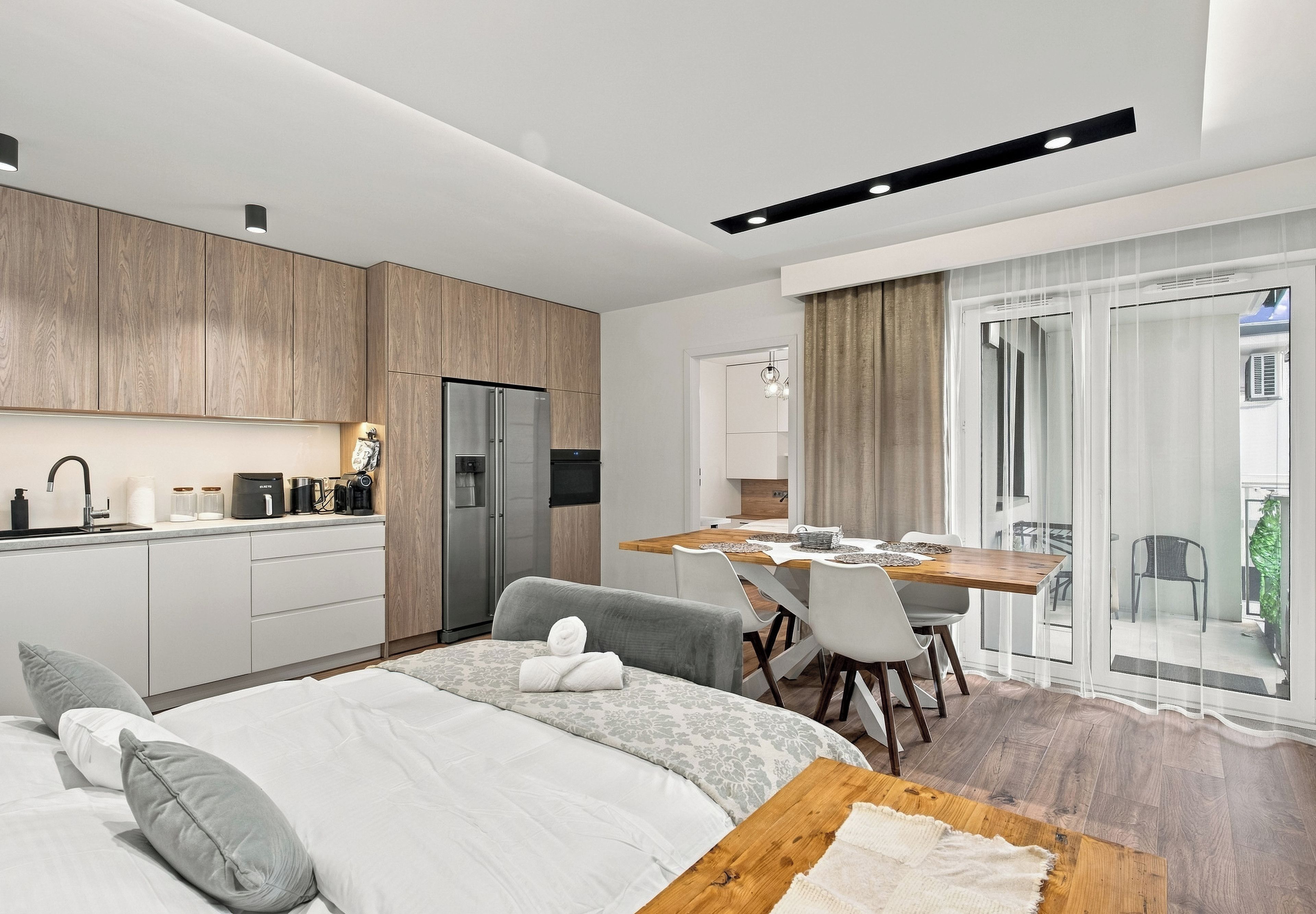 Une chambre d’hôtel moderne dotée d’une kitchenette, d’un coin repas, d’un lit avec un canapé gris et d’un balcon avec des rideaux.