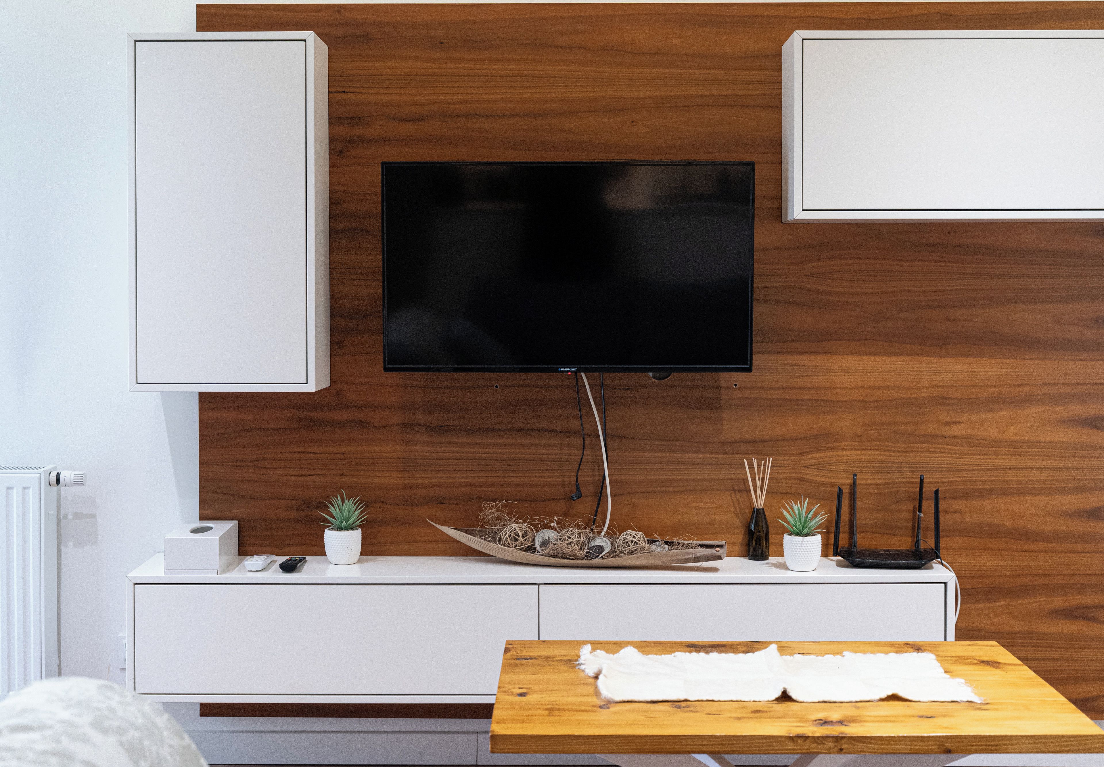 Ein modernes Wohnzimmer mit einem flachbildfernseher an einer Holzwand, einer weißen Unterhaltungseinheit darunter und einem Holztisch mit weißem Tuch.