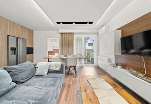 Ein moderner Wohnraum mit einer Couch, einem Esstisch, einer Küchenzeile und einem an der Wand befestigten Fernseher.
